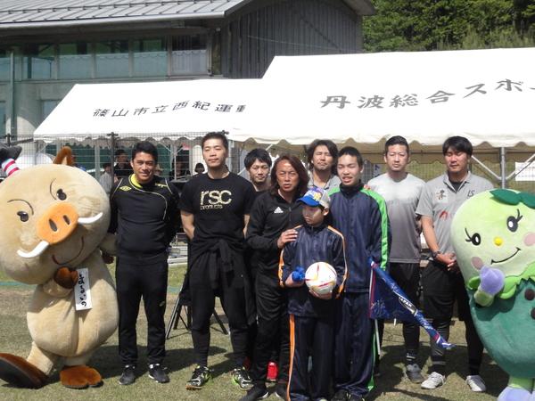 サッカーボールを持っている男の子の肩に手を置いている北澤 豪さんの後ろで他のサッカー選手と選手の脇に丹波篠山キャラクター（まるいの、まめりん）が写っている写真