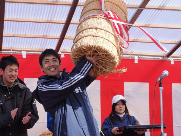男性が赤白のテープが結ばれている米俵を高く持ち上げ笑顔で喜んでいる写真