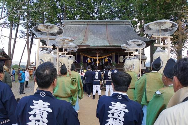和傘の下に提灯が下げられているものを持っている神主さんたちと法被を着た人々が神社に向かって並んでいる写真