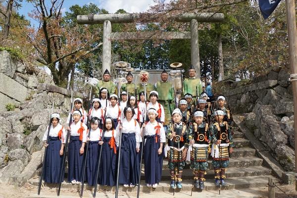 神社の鳥居前の階段で鉢巻きに袴姿で槍を持っている女の子らと鉢巻きに鎧姿で槍を持っている男の子らと神主さんらが一緒に記念撮影している写真