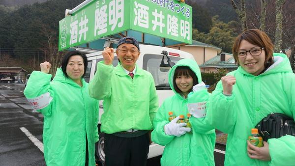 選挙カーの前で市長と女性3人が黄緑色のジャンパーを着て、右手を上げ笑顔で記念撮影している写真