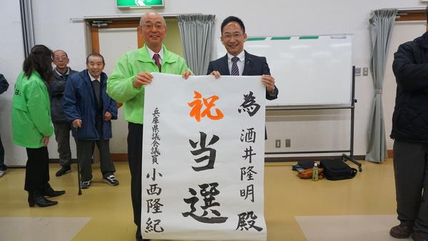 小西県議より大きな模造紙に「祝 当選」と書かれた紙をお祝いでいただき、手に取りながら市長と小西県議が記念撮影している写真