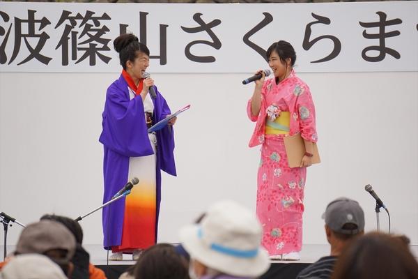 ステージ上で白地に下が黄色とオレンジのグラデーションの色を着て上に紫色の羽織を着ている女性と右横にピンク地の花柄の着物を着ている女性が楽しそうに話しをしている写真