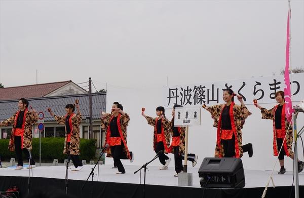 ステージ上で黒地に金色をベースにした花柄の法被を着た「弥華美」チームが鳴子を両手に持って左足を後ろに上げて踊っている写真