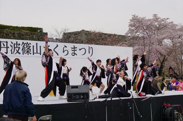 ステージ上で右袖のみ着物の花柄が入っている黒色の法被を着た男女混合のチーム「輝楽」のメンバーが両腕を左右それぞれに思いっきり腕を伸ばしてポーズを決めている写真
