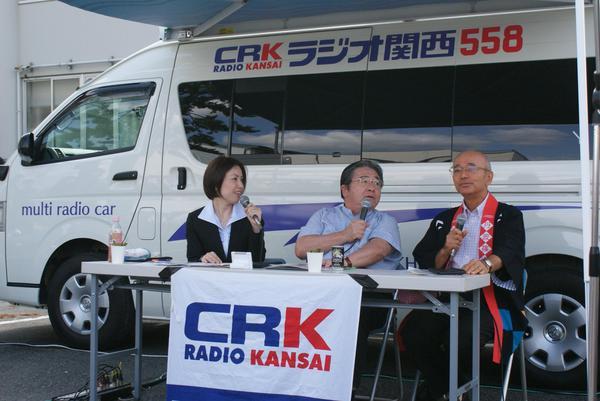 田辺眞人のまっことラジオの生放送でラジオカーの前に机と椅子を置いて座り、女性アナウンサーと眞人アナウンサーと市長がマイクで話をしている写真