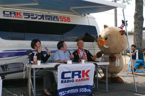 田辺眞人のまっことラジオの生放送で女性アナウンサーと眞人アナウンサーと市長がマイクで話をしていて、マスコット「まるいの」が参加している写真