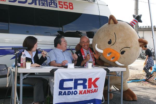 田辺眞人のまっことラジオの生放送で女性アナウンサーと眞人アナウンサーと市長がマイクで話をしていて、マスコット「まるいの」が市長にハグしている写真