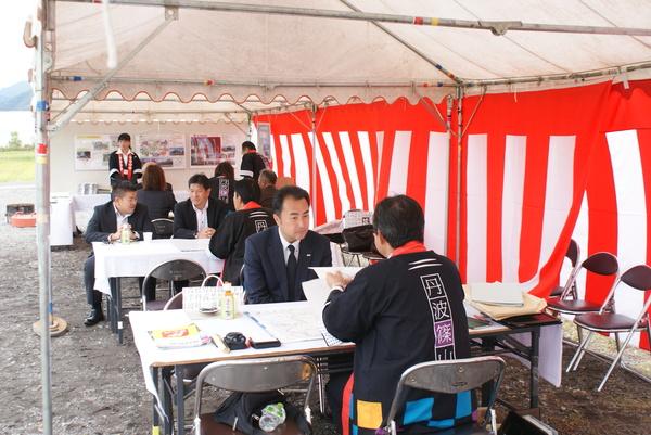 法被を着た男性がテントに準備された机で見学者に話をしている写真