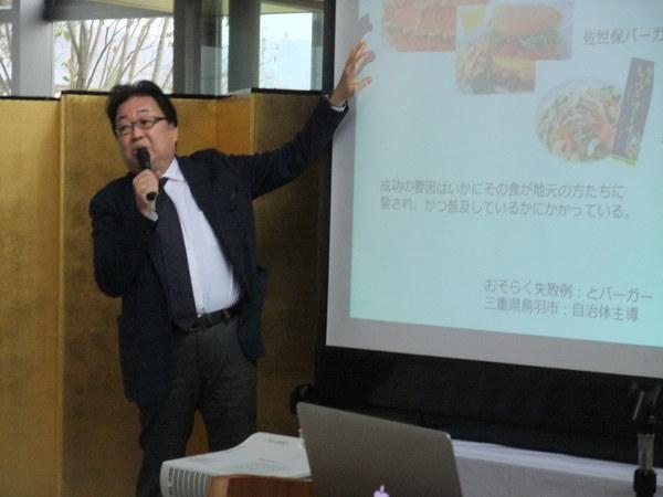 スクリーンに映像を映しながら、青山 忠靖さんがマイクを持って講演している様子の写真