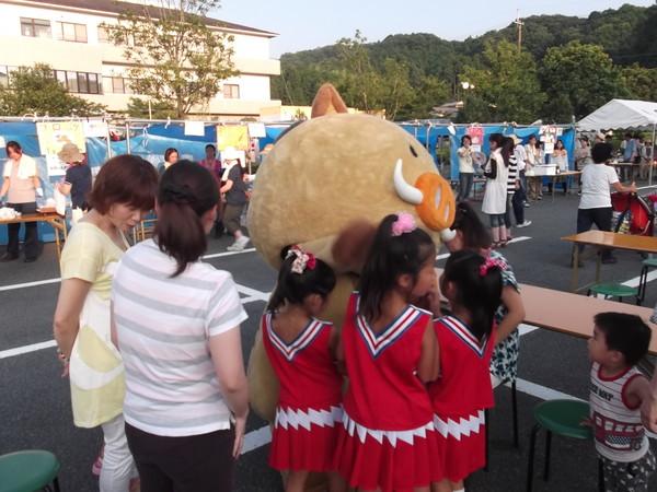 ゆるキャラの「まるいの」の周りに赤いユニホームを着た女の子3人と小さい男の子が集まって来ており、後ろの方ではお祭りの屋台が準備されている様子の写真