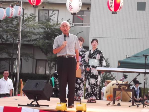 市長が舞台の上でマイクで話をしており、後には浴衣を着た女性二人が立っている様子の写真