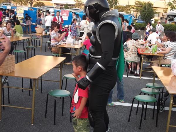 ニューヒーロー「ササグロー」と赤いティーシャツを着た男の子が記念写真を撮っている様子を横から写しており、周りには家族連れで遊びに来ているお客さんがテーブルに座って飲食を楽しんでいる様子の写真