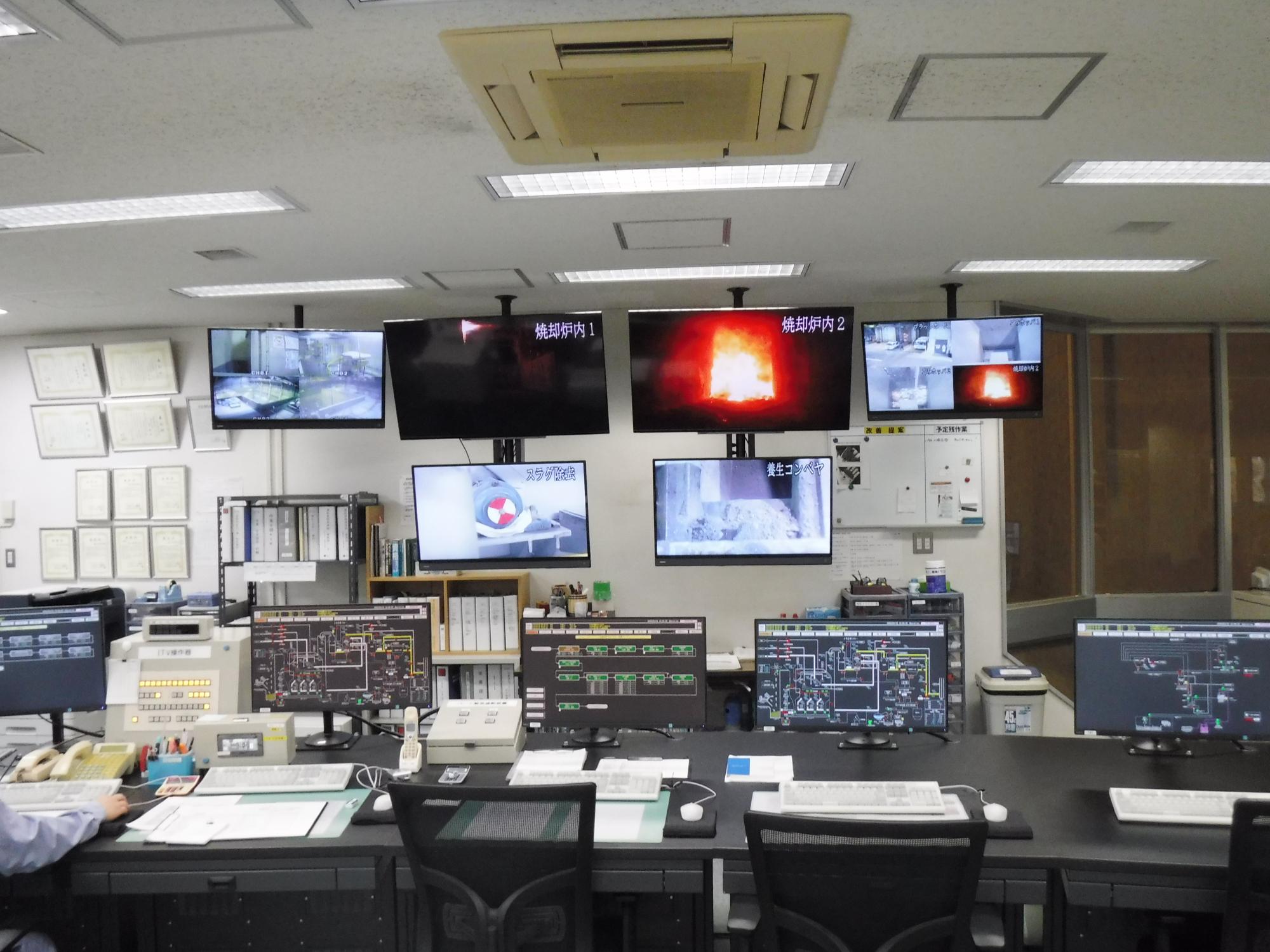 事務所にたくさんのパソコンやモニターがあり、大きめのモニターが2つの右側は火が燃えている様子が映し出されている写真