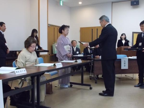 紫の着物の女性がスーツを着た男性から表彰をされている写真