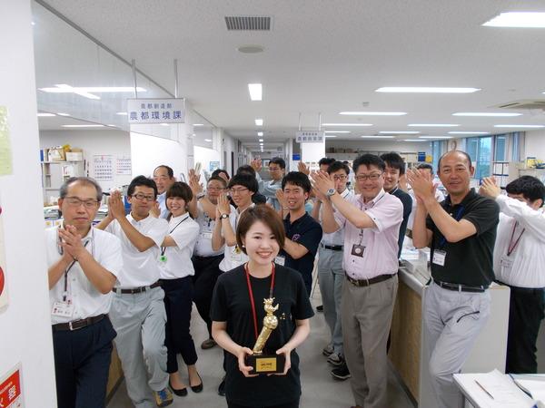 チャンピオンになった小林 真弓さんがトロフィーを持ち、その後方に両手を顔の右横に置き、笑顔で拍手をしている社員の方々の写真