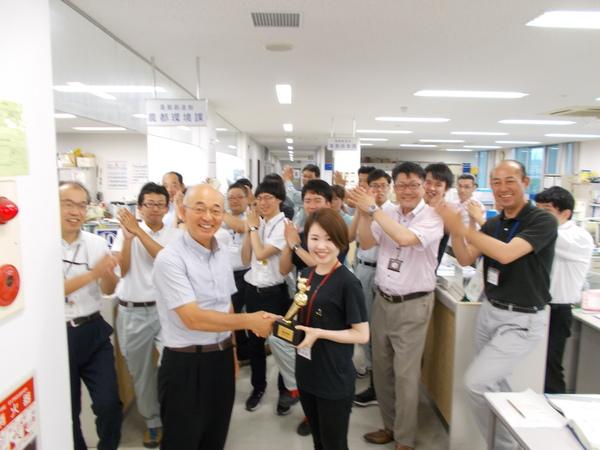 市長と小林 真弓さんが笑顔で握手をし、その後方で拍手をしている社員の方々の写真