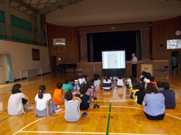 体育館の中で子供たちが体育座りをしてスクリーンを使って説明している市長の話を聞いている写真