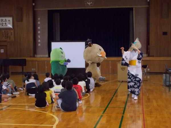 浴衣を着た方とマスコットキャラクターが体育館で座っている子供達の前を踊りながら通りすぎている写真