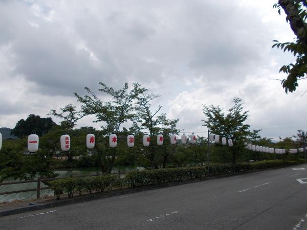 奥には池がある道路沿いに岸本設備と書かれた提灯が飾られている写真