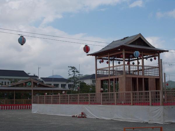 広場に木で作られたヤグラが建てられていて、手前に提灯が飾られている写真