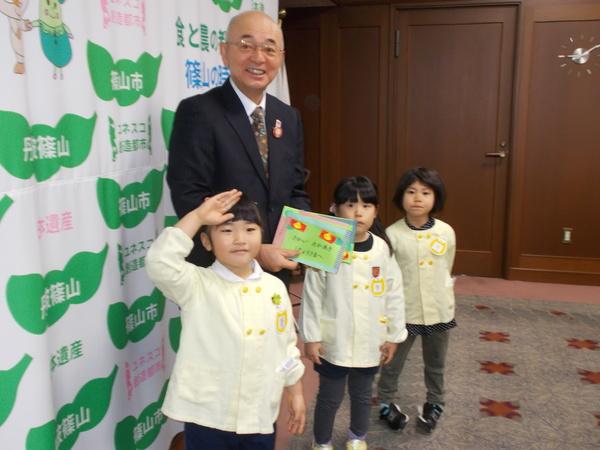 市長が「さかい たかあきしちょうさまへ」と書かれた招待状を手に持っており、黄色いスモックをきた園児と一緒に写っている写真