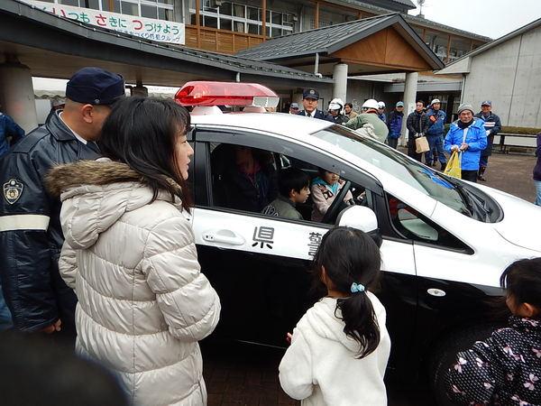 パトカーの中に男の子と女の子が乗って中を興味深く見ていてその様子を近くで警察官が見守っている写真