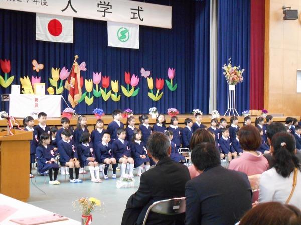 ステージに入学式と書かれた幕と、カーテンには赤色、黄色、ピンク色の紙で作られたチューリップが貼られており、学校の制服を着た新入生がステージを背に保護者と向き合って椅子に座っている写真