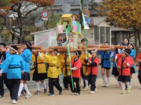 青色、黄色、赤色の法被を着た子ども達が酒樽を飾り付けたおみこしを担いで通りを歩いている様子の写真