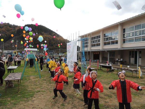 学校の校庭で沢山の参加者が、空に色とりどりの風船を飛ばし、手前にいる法被を着た子ども達が、空に上がっていく風船を嬉しそうに見上げている様子の写真