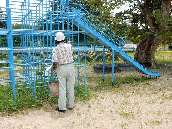 ヘルメットをかぶった男性が青色のジャングルジムを握っており、遊具の点検をしている様子の写真