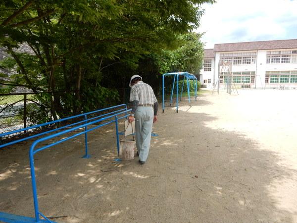 ヘルメットをかぶった男性が運動場に設置されている青色の平行棒を見ながら、遊具の点検をしている様子の写真