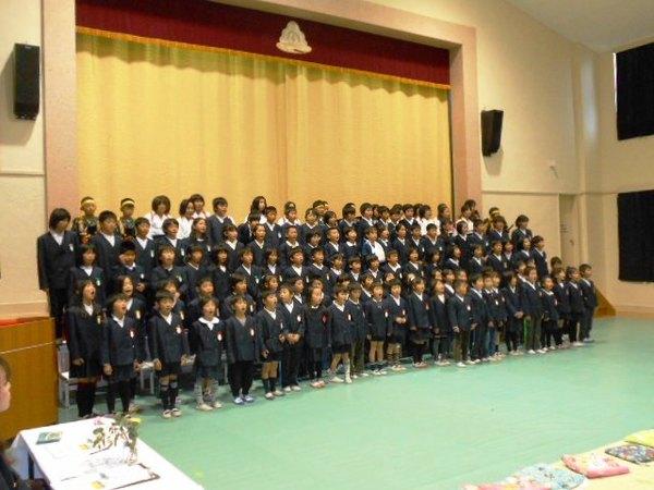 制服を着た児童たちが舞台の前に段々に並んで顔が見えるように立ち、合唱をしている写真