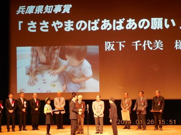 舞台上のスクリーンに兵庫県知事賞「ささやまのばあばあの願い」と写しだされ、その前で 阪下 千代美様が受賞されている写真