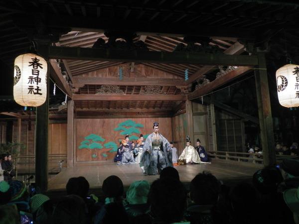 春日神社の境内で3名のこづつみと1名の笛の演奏をバックに男性が三番叟の薄い青色の衣装を着て両手を広げ元朝能の舞を披露している写真