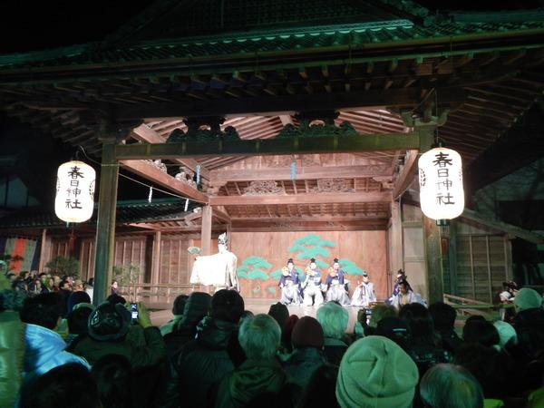 春日神社の境内で3名のこづつみと1名の笛の演奏をバックに三番叟の金色の衣装を着た男性が扇子を右手に持ち元朝能の舞を披露している写真