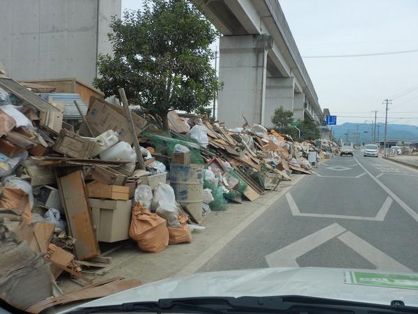 道路際に災害で出た机や木片等、高くつみあげられた廃棄物が遠くまで続いている様子の写真