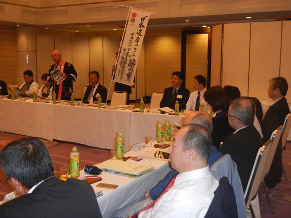 全国京都会議の会議で、「日本遺産の町 篠山市」と書かれているのぼりを男性が持ち、市長が資料をもって話をしており、周りの人は資料に目を通しながら話を聞いている人や市長の話を聞き入っている様子の写真