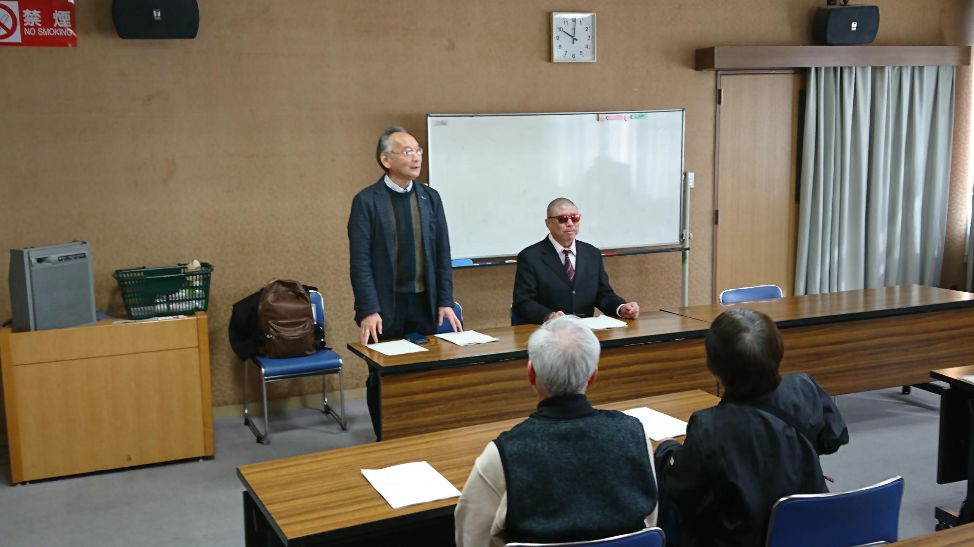 前に座っている男性が2人おり、左側の男性が立って会場内に向けて話している写真