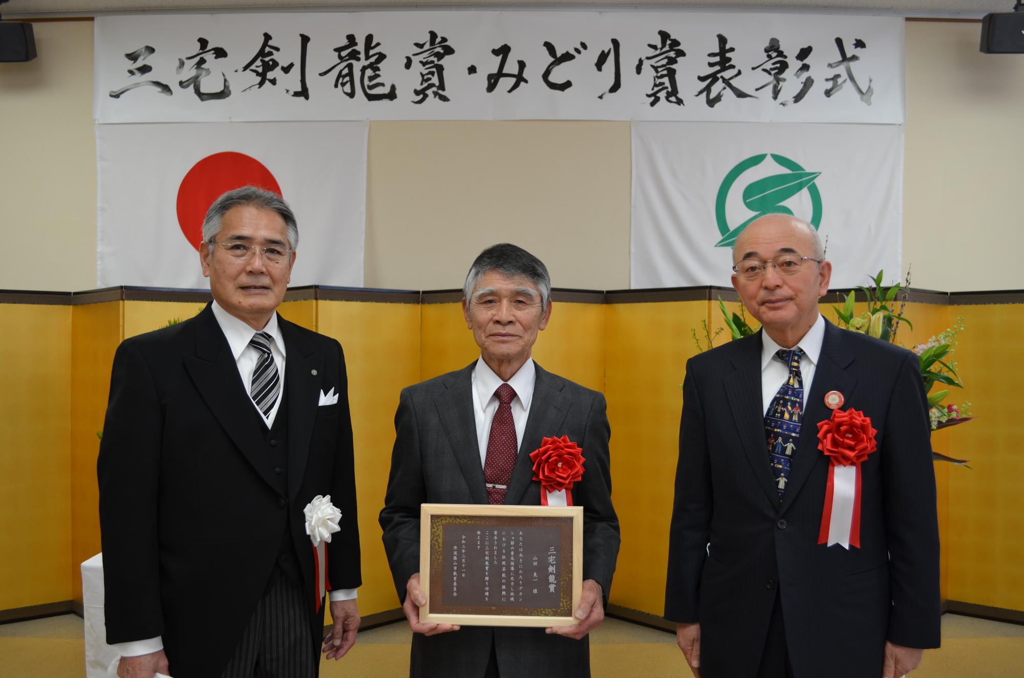 教育長と市長、表彰盾を手に持つ山田さん。