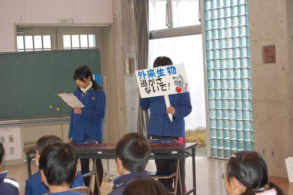 外来生物逃がさないで！と書かれた立て札を持って、女子生徒2名が発表している写真