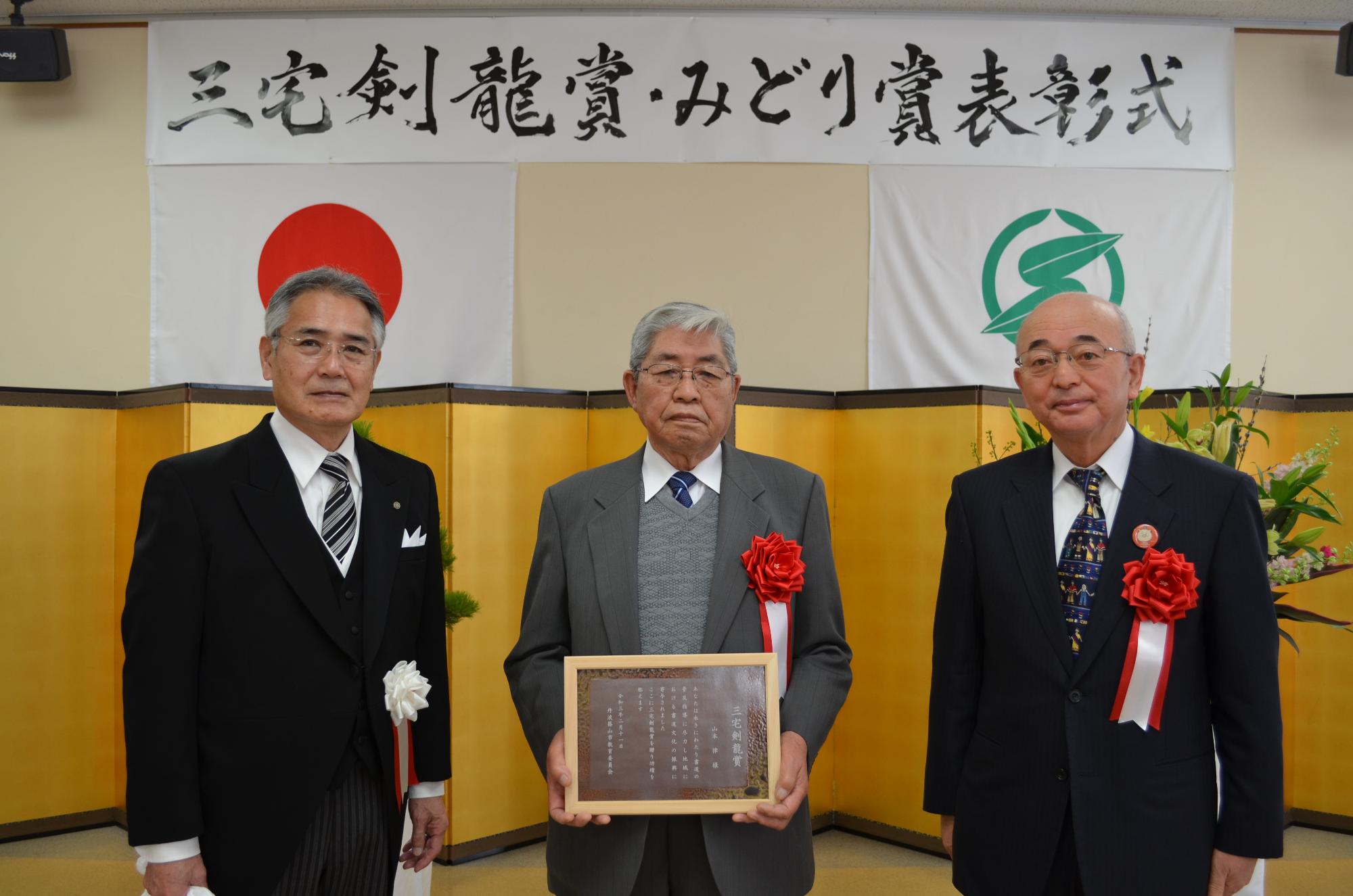 教育長と市長、表彰盾を手に持つ山本さん。