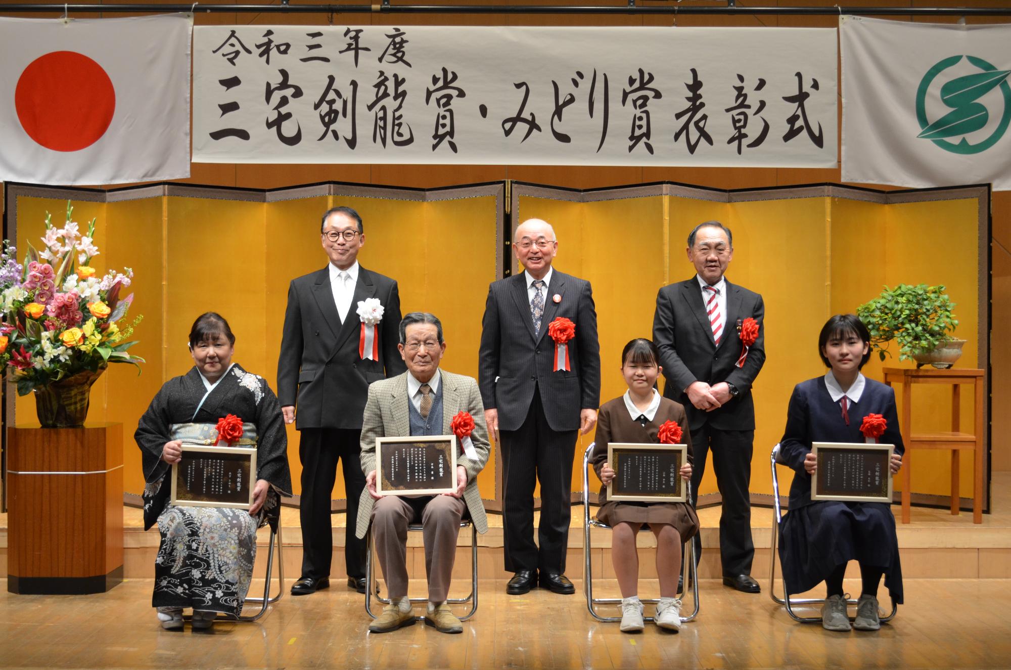 受賞者4名が前列に座ってひざに表彰盾をかかげている。後列に市長、教育長、議長が立っている