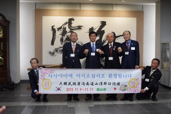 ロビーに掲げられた書の前で市長と韓国郡守のホギドさんらが握手をして横に並び、その前に座って日韓の横断幕を2名の男性職員が支えている写真
