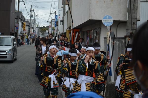 狭い道を鎧を羽織った子供達が歩き横をグレーの軽自動車が走っている写真