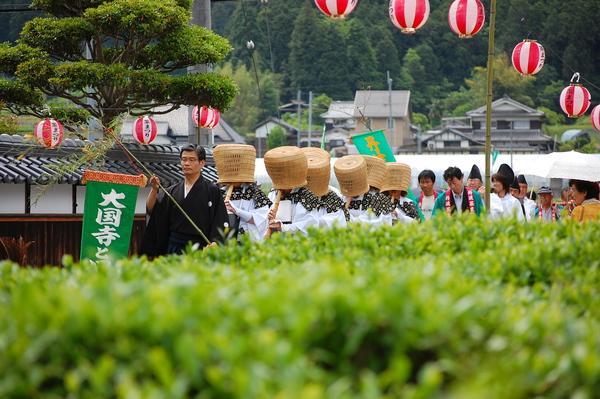 沿道に提灯が飾られ、黒の紋付袴を着た男性が大国寺と書かれた緑色の幕を持ち、その後ろに白い衣装に天蓋の帽子を被り、木の笛を吹いた人々が続き、その後方には法被を着た方々が一列に並んで歩いている写真