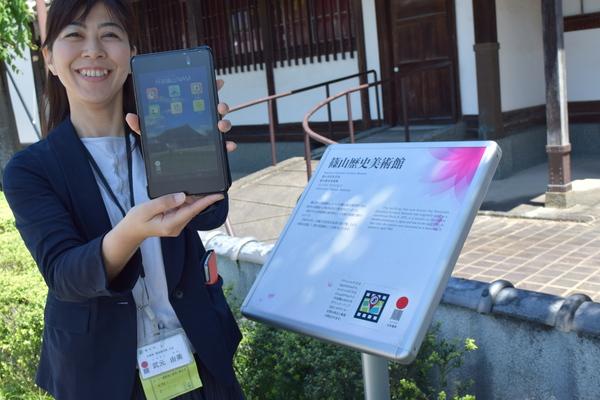 歴史美術館前に設置された看板の前に女性が笑顔でスマートフォンを持って見せている写真