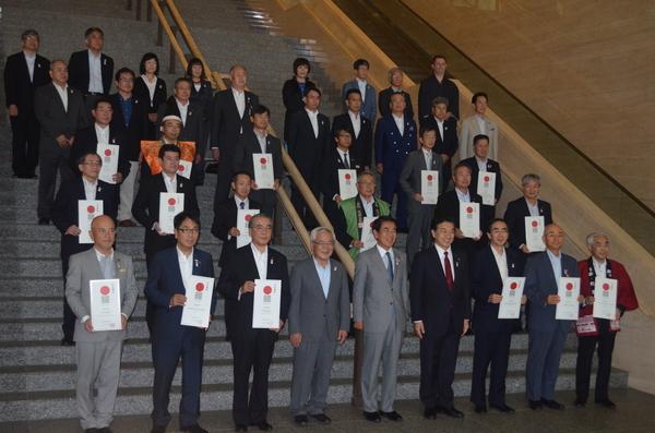 スーツを着た人たちや法被を羽織った方々が認定書を手に持ち階段の所でみんなで記念写真