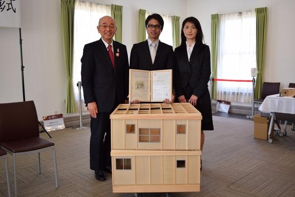 住宅の模型の後ろで賞状の入った額を持った受賞者の夫婦が市長と一緒に記念撮影している写真