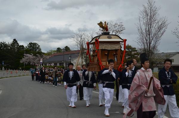 ピンク色の装束を着た男性の後ろをすぐ、神輿を担いでいる人たちの写真